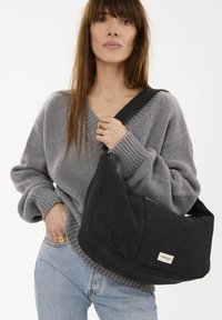 Sac à épaule en tissu noir avec une forme unique, présentant une fermeture éclair visible et un patch logo ; associé à un pull en tricot gris et un jean bleu.