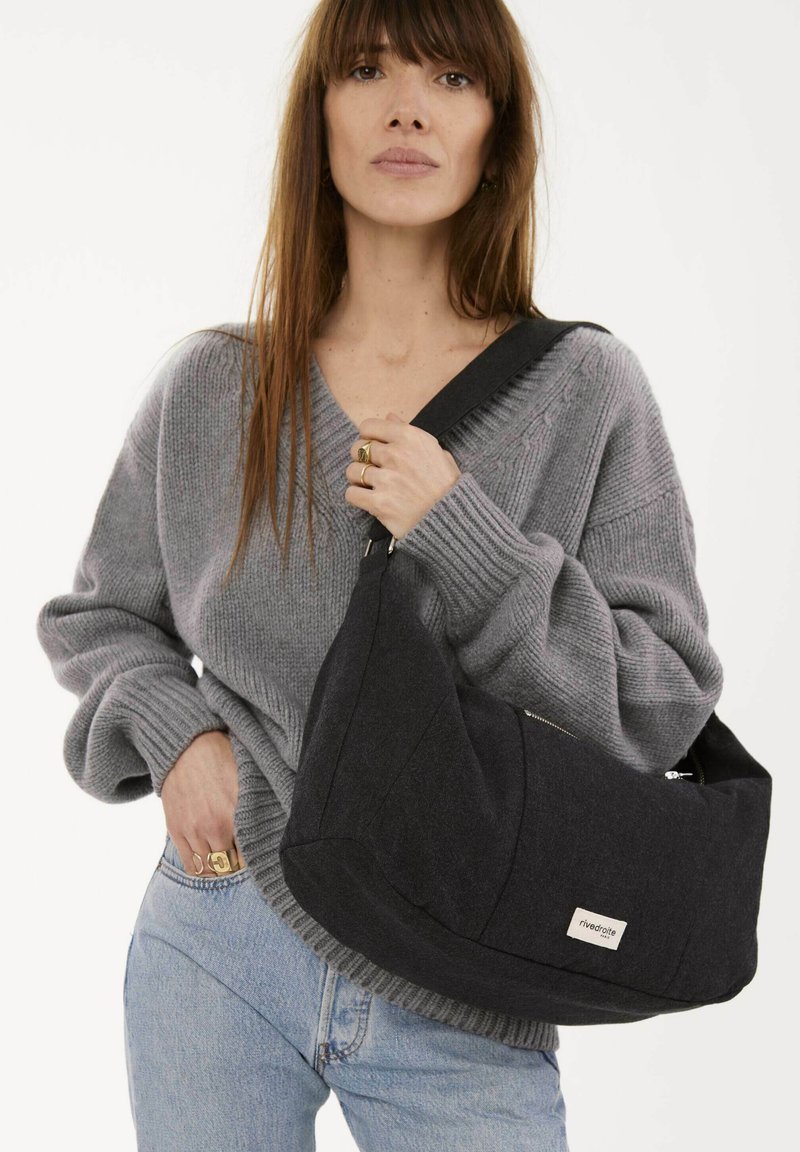 Sac à épaule en tissu noir avec une forme unique, présentant une fermeture éclair visible et un patch logo ; associé à un pull en tricot gris et un jean bleu.