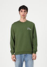 Grüner Baumwoll-Sweatshirt mit langen Ärmeln, Rundhalsausschnitt und gerippten Bündchen. Mit weißem Logodetailing auf der linken Brust. Trägt eine blaue Jeans.