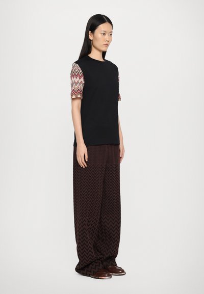 Μαύρη μπλούζα T-shirt με κανονικά μανίκια με σχέδιο, συνδυασμένη με παντελόνι φα�ρδύ με σχέδιο ζιγκ ζαγκ σε καφέ απόχρωση. Το μοντέλο στέκεται σε ουδέτερο φόντο.
