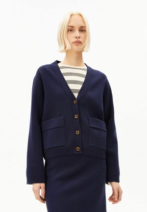 Junge Frau trägt eine marineblaue, geknöpfte Strickjacke über einem gestreiften Top und einem passenden marineblauen Rock, sie steht vor einem schlichten weißen Hintergrund.