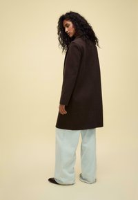 Long brown coat made of soft fabric, featuring a straight design and notched collar, worn over light blue wide-leg jeans.