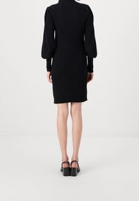 Robe en maille côtelée noire avec des manches bouffantes, silhouette ajustée et longueur midi. Associée à des chaussures à talons blocs noires.