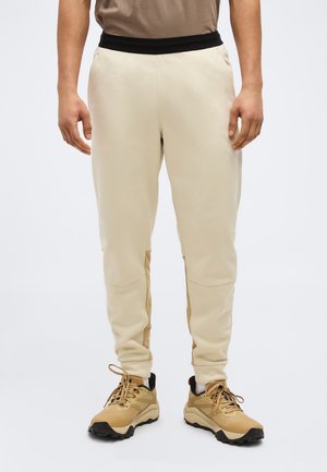 Man die beige taps toelopende broek draagt met een zwarte tailleband en tan wandel Schoenen, staand tegen een effen lichte achtergrond.
