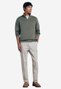 Maglione verde oliva chiaro con colletto zip, abbinato a pantaloni cargo beige con tasche laterali. Il modello indossa scarpe slip-on marroni.
