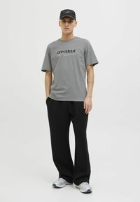 Grå T-shirt med korte ærmer og sort logo-tekst. Sorte vide bukser. Sølv sneakers med hvide accenter. En sort kasket fuldender outfittet.