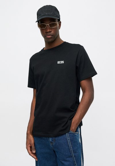 Čierne bavlnené tričko s krátkymi rukávmi s bielym logom "GCDS" na hrudníku. Model ho kombinuje s modrými džínami a čiernou čiapkou.