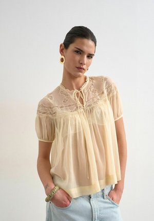 Femme aux cheveux foncés portant un chemisier en dentelle beige transparente, un jean bleu clair, de grandes boucles d'oreilles rondes et un bracelet rayé, posant devant un fond uni.