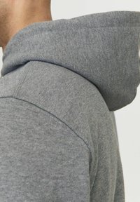 Felpa grigia con una texture morbida, dotata di un cappuccio arrotondato e cuciture visibili lungo la cucitura della spalla. Ingrandimento del dettaglio del colletto.