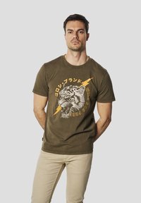 Giovane uomo che indossa una maglietta verde oliva con grafica di una tigre ruggente e un fulmine giallo, abbinata a pantaloni beige, in piedi davanti a uno sfondo grigio.