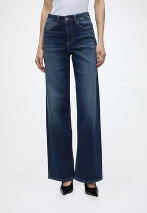 Femme portant un jean bleu foncé à jambes larges et des chaussures noires à bouts pointus, montrée de la taille aux pieds sur un fond blanc.