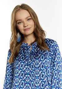 Blusa blu con motivo e scollo a cravatta, caratterizzata da una combinazione di tonalità blu, teal e bianco in un tessuto fluido e morbido.