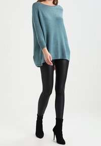 Luźny, zielonkawo-niebieski sweter z okrągłym dekoltem i opuszczonymi ramionami, zestawiony z czarnymi, błyszczącymi legginsami oraz czarnymi szpilkami na kostce.