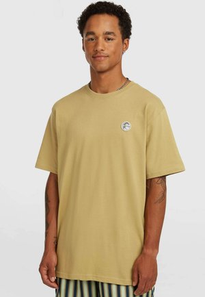 T-shirt beige in cotone con maniche corte, scollo rotondo e un piccolo logo rotondo sul petto a sinistra. Modello che indossa pantaloni a righe.