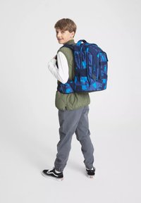 Zaino camouflage blu, spallacci imbottiti, molteplici tasche, forma rettangolare, tessuto resistente. Indossato da un bambino con abbigliamento chiaro.