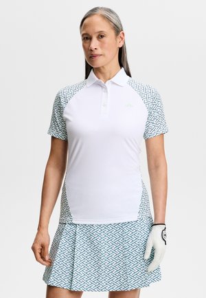 Kobieta ubrana w białą rękawiczkę golfową, białą koszulkę polo z wzorzystymi rękawami oraz pasującą, wzorzystą spódnicę, stojąca na jednolitym tle.