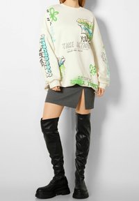 Sudadera blanca de gran tamaño con estampados gráficos en verde y negro, combinada con una falda gris con una abertura lateral. Con botas negras hasta la rodilla.