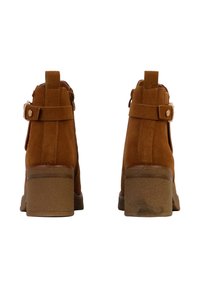 Bottes en suède tan à la cheville avec un talon bloc de hauteur intermédiaire, présentant une sangle et un détail de bouton à la cheville, et une semelle légèrement usée.