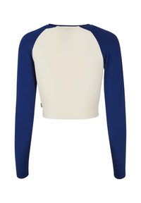 Lange mouwen cropped top in crème en marineblauw. Heeft raglanmouwen, een zachte textuur en contrasterende stiksels langs de schoudernaden.