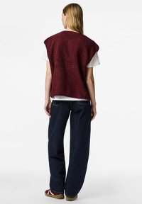 Vinröd ärmlös tröja med texturerat material, kombinerad med en vit t-shirt och mörkblå jeans med vida ben. Platta skor fullbordar outfiten.