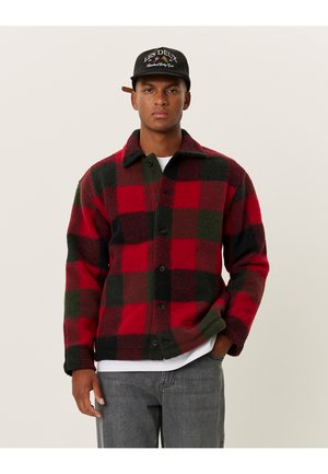 Red and green checkered flannel jacket with a button front, collar, and long sleeves, paired with gray pants and a black cap.
