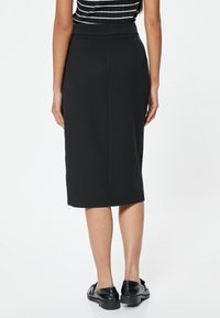 Zwarte kokerrok met een gladde textuur, knie-lange snit, elastische tailleband en een eenvoudige uitstraling. Draagt met zwarte platte schoenen.