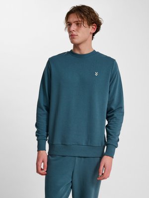 Junger Mann mit lockigem Haar, der einen türkisfarbenen Rundhalspullover und passende Hosen trägt, schaut nach links vor einem einfarbigen grauen Hintergrund.