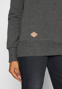 Persoon draagt een donkergrijze sweater met witte stippen, een zandkleurig logo-patch en donkerblauwe spijkerbroek, hand rustend naast het dijbeen.