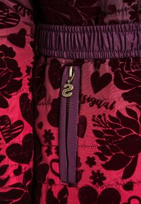 Chaqueta acolchada con un estampado floral en rosa y burdeos. Presenta una cinturilla elástica morada y un cierre metálico con un tirador con logotipo.