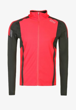 Homme marchant vers l'avant portant une veste zippée rouge et noire, un t-shirt de sport noir et un short, avec des chaussures de course blanches et orange.