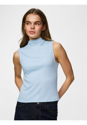 Ung kvinde med langt hår iført lyseblå ærmeløs mock-neck top og mørke jeans, ser til siden med hænderne bag ryggen.