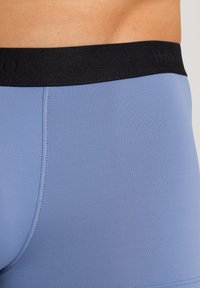 Blå boxerkalsonger med svart elastisk midjeband. Slät textur och åtsittande design, med sömmar längs sidorna för extra detalj.