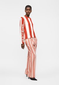 Γυναίκα που φοράει κόκκινο και άσπρο ριγωτό πουλόβερ με κείμενο στο μανίκι και φαρδιά ριγωτά παντελόνια σε κοραλί και άσπρο.