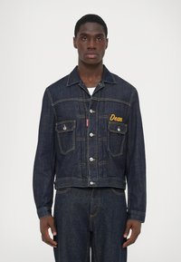 Chaqueta de mezclilla oscura con un ajuste corto, cierre de botones, bolsillos en el pecho y nombre bordado en amarillo "Dean". Presenta costuras en contraste y herrajes plateados.