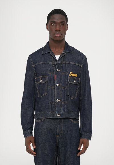 Mørk denimjakke med cropped pasform, knaplukning, brystlommer og gult broderet navn "Dean." Har kontrasterende syninger og sølvhardware.
