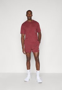 Camiseta de manga corta color marrón oscuro con un bolsillo de parche, combinada con pantalones cortos a juego. El material tiene un aspecto texturizado. Zapatillas blancas y calcetines completan el conjunto.