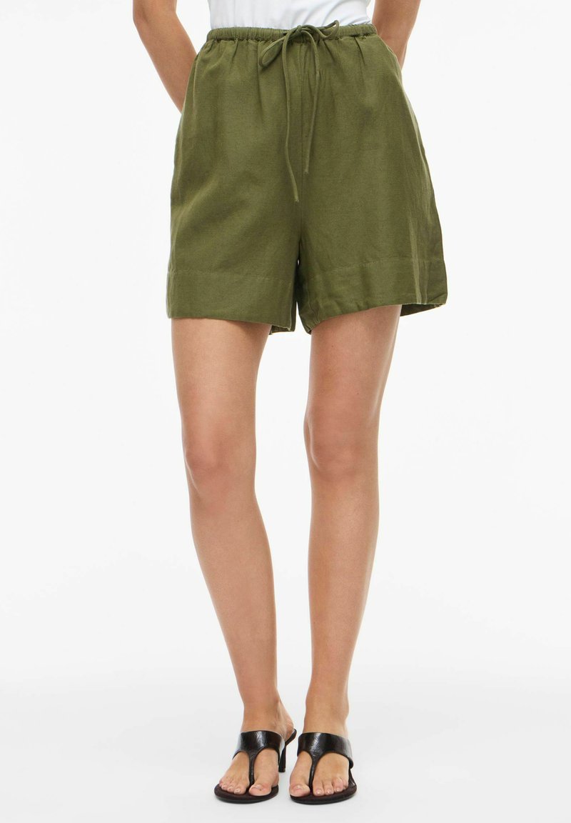 Femme portant un short en lin vert olive à cordon et des sandales à talons noires, debout les mains dans le dos.