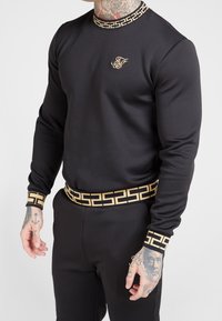 Svart atletisk sweatshirt med guldaccenter. Har ett mönstrat krage och manschetter, en snug passform och matchande midja.