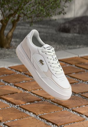 Witte vetersneaker van glad leer en suède, met een gouden krokodillenlogo en een gestructureerde rubberen zool, gepositioneerd boven houten tegels.