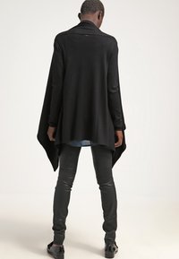 Černý lehký kardigan s volným střihem, dlouhými rukávy a lemem, který se na bocích zakřivuje, nošený přes přiléhavé černé kalhoty.