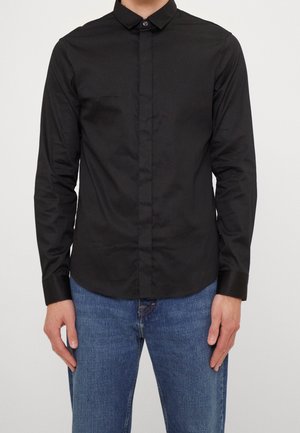 Homme portant une chemise noire ajustée à manches longues avec patte de boutonnage cachée et un jean bleu, debout devant un fond uni.