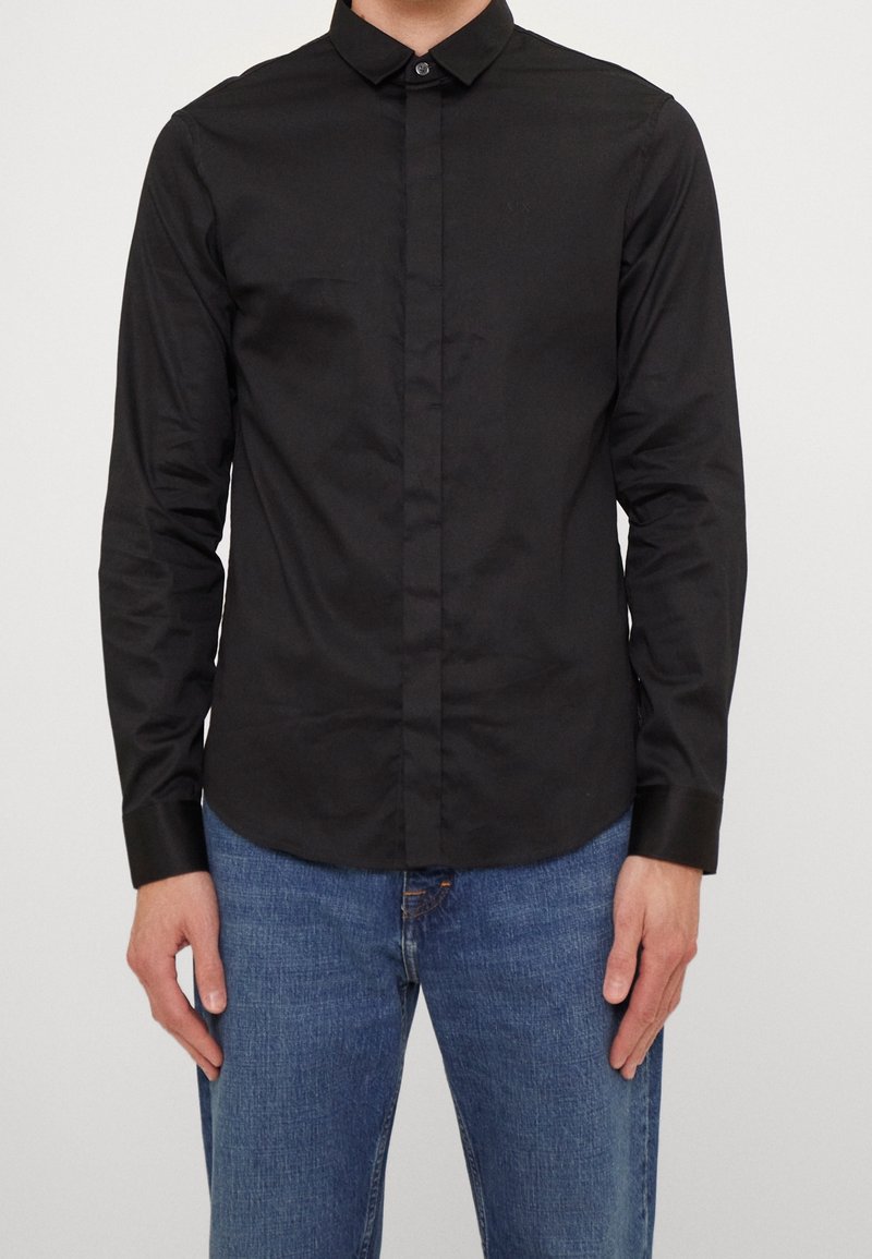 Homme portant une chemise noire ajustée à manches longues avec patte de boutonnage cachée et un jean bleu, debout devant un fond uni.