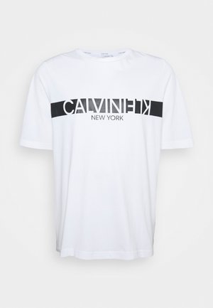 Camiseta blanca de algodón con una franja horizontal negra y la frase "CALVIN KLEIN NEW YORK" impresa en letras en negrita. Cuello redondo, mangas cortas.