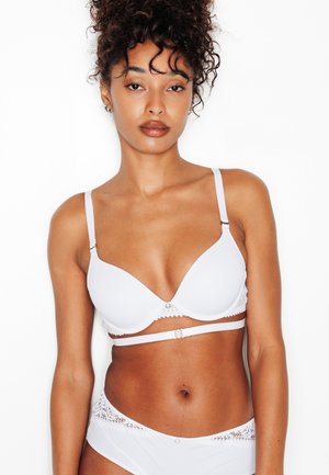 Femme aux cheveux bouclés portant un soutien-gorge blanc et un sous-vêtement assorti orné de dentelle blanche, debout devant un fond blanc uni.