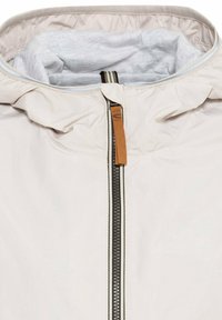 Veste à capuche beige clair avec une fermeture éclair noire et blanche et un passant en tissu marron, montrant une doublure grise douce à l'intérieur de la capuche.