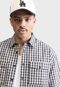 Schwarz-weiße kariertes Flanellhemd mit zwei Brusttaschen, Knopfverschluss, über einem weißen Tanktop getragen, ergänzt durch eine weiße Kappe.