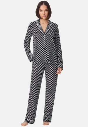 Femme debout pieds nus, portant un haut de pyjama à manches longues boutonné à motifs noirs et blancs, assorti d'un pantalon avec un galon blanc.