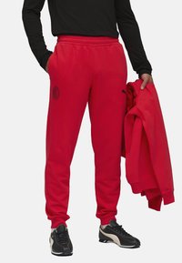 Røde sweatpants med elastisk talje, der har et sort logo. Modellen har en sort langærmet trøje på og holder en matchende rød hættetrøje. Sorte sneakers.