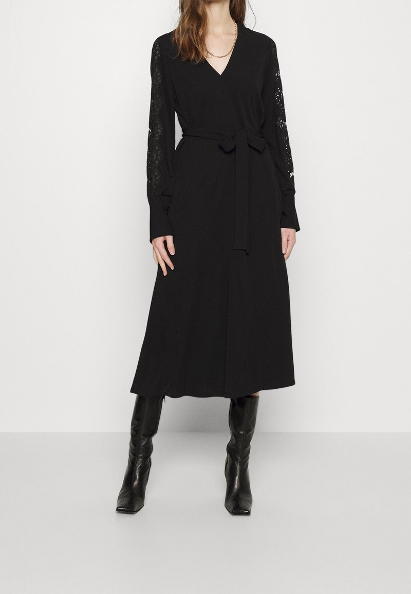 Femme portant une robe midi noire à manches longues en dentelle, une ceinture à la taille, et des bottes en cuir noires montantes jusqu'aux genoux, debout sur un fond blanc.