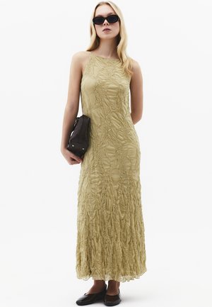 Kobieta w beżowej, bezrękawnikowej, teksturowanej sukience maxi, czarnych balerinach, czarnej kopertówce i ciemnych owalnych okularach przeciwsłonecznych, stojąca na białym tle.